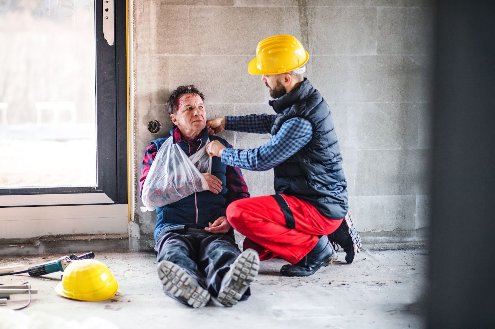 A construction worker wearing a navy vest and red work pants kneels beside an injured colleague with a bandaged arm and head wound, providing aid in a partially constructed building with tools and a yellow hard hat scattered on the floor.